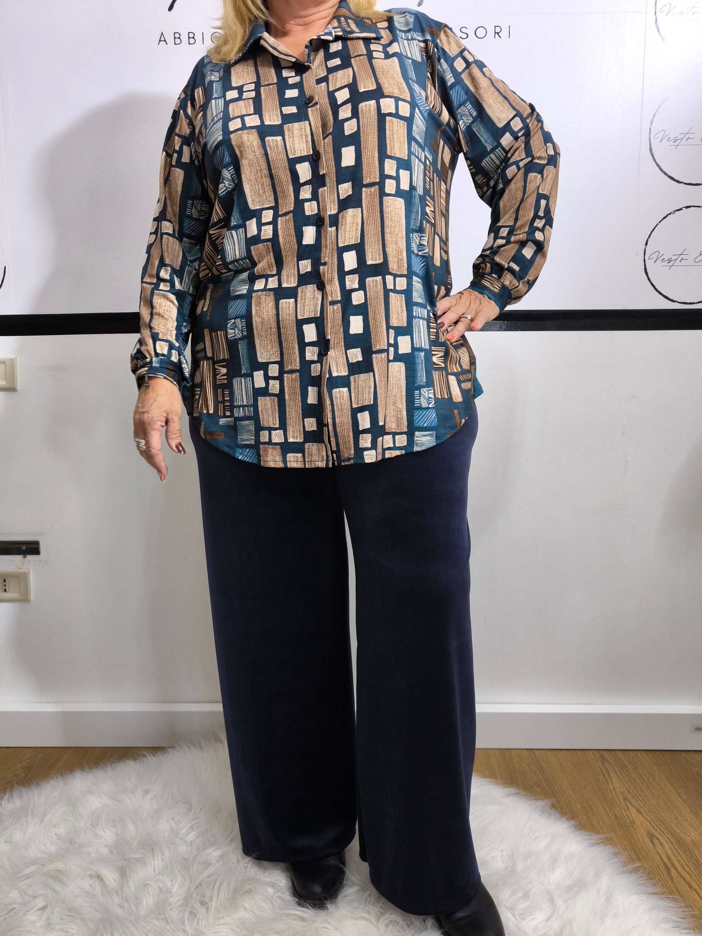 Donna che indossa una camicia in viscosa blu petrolio con stampa geometrica, affiancata dalla stessa camicia in tonalità nera e dorata su appendino.