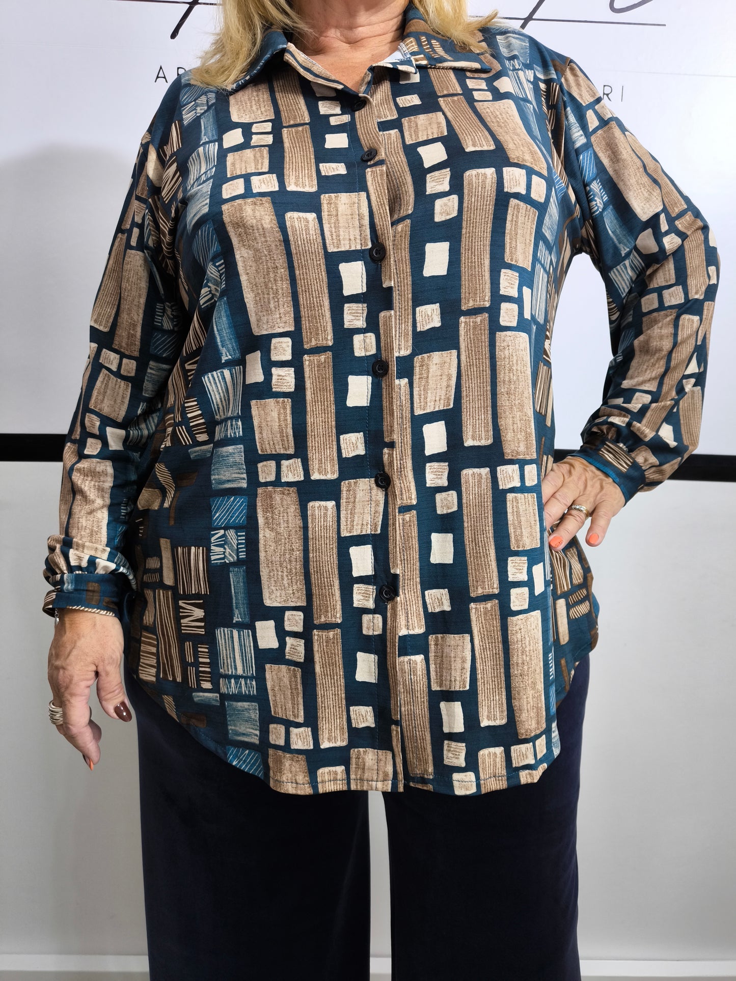 Donna che indossa una camicia in viscosa blu petrolio con stampa geometrica, affiancata dalla stessa camicia in tonalità nera e dorata su appendino.