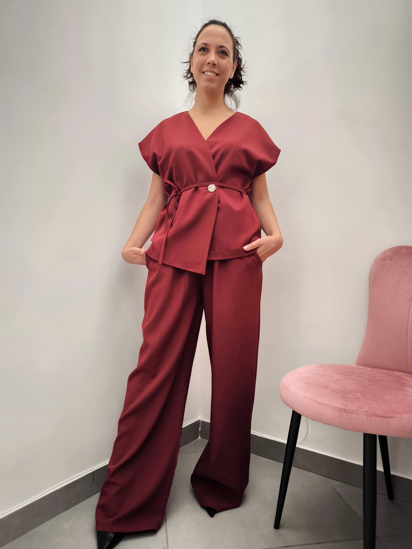 Donna che indossa completo elegante AURA in tessuto bordeaux, con giacca foderata a manica corta e pantaloni larghi.