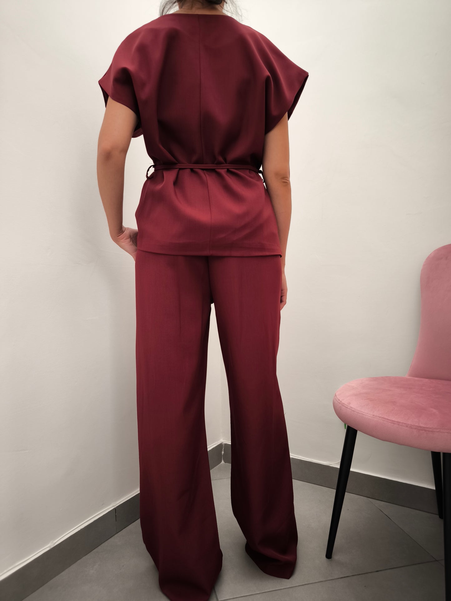 Donna che indossa completo elegante AURA in tessuto bordeaux, con giacca foderata a manica corta e pantaloni larghi.