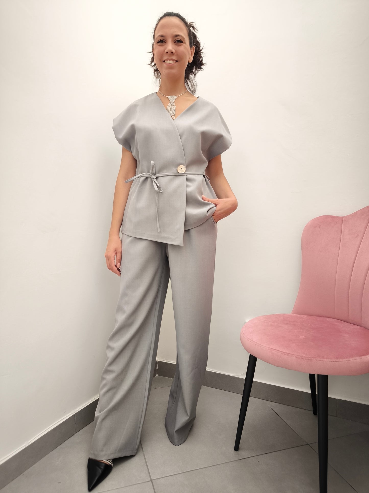 Donna che indossa completo elegante AURA in tessuto grigio, con giacca foderata a manica corta e pantaloni larghi.