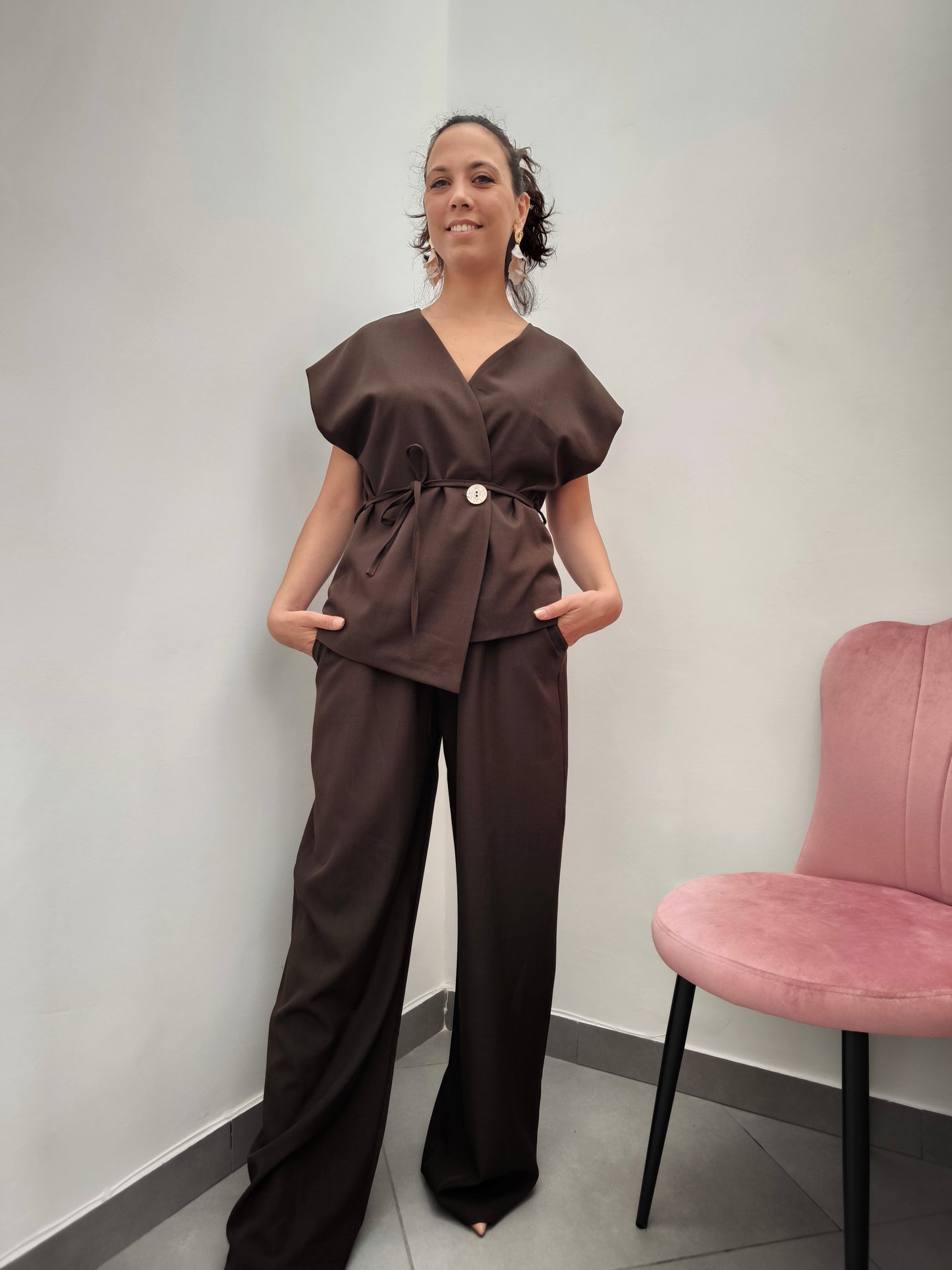 Donna che indossa completo elegante AURA in tessuto marrone, con giacca foderata a manica corta e pantaloni larghi.