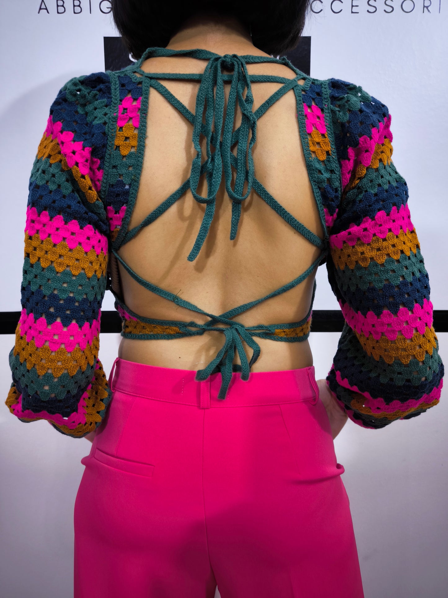 Maglia crochet multicolor a righe con schiena incrociata, abbinata a pantaloni fucsia e cintura dorata, indossata da una modella sorridente.