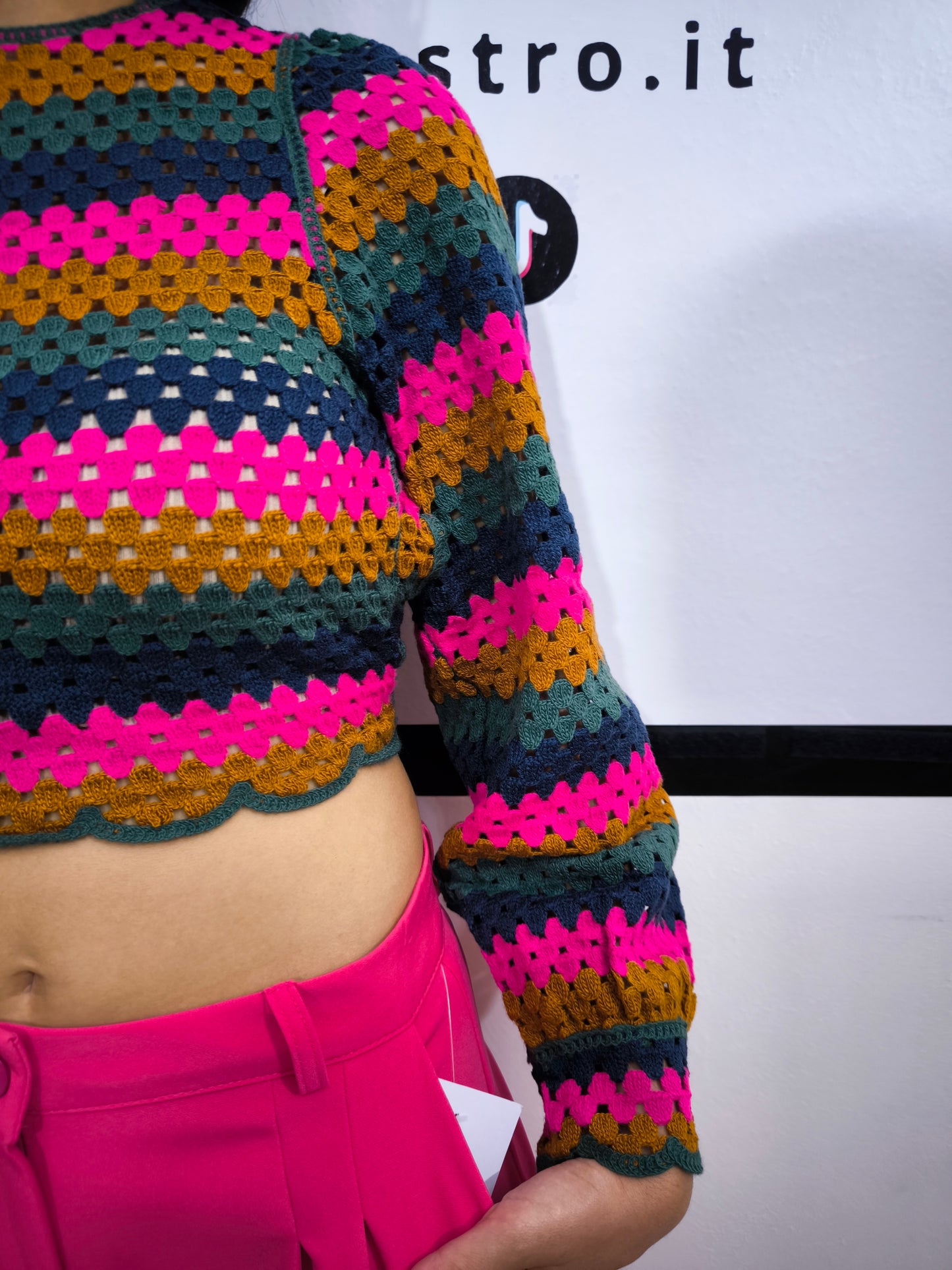 Maglia crochet multicolor a righe con schiena incrociata, abbinata a pantaloni fucsia e cintura dorata, indossata da una modella sorridente.