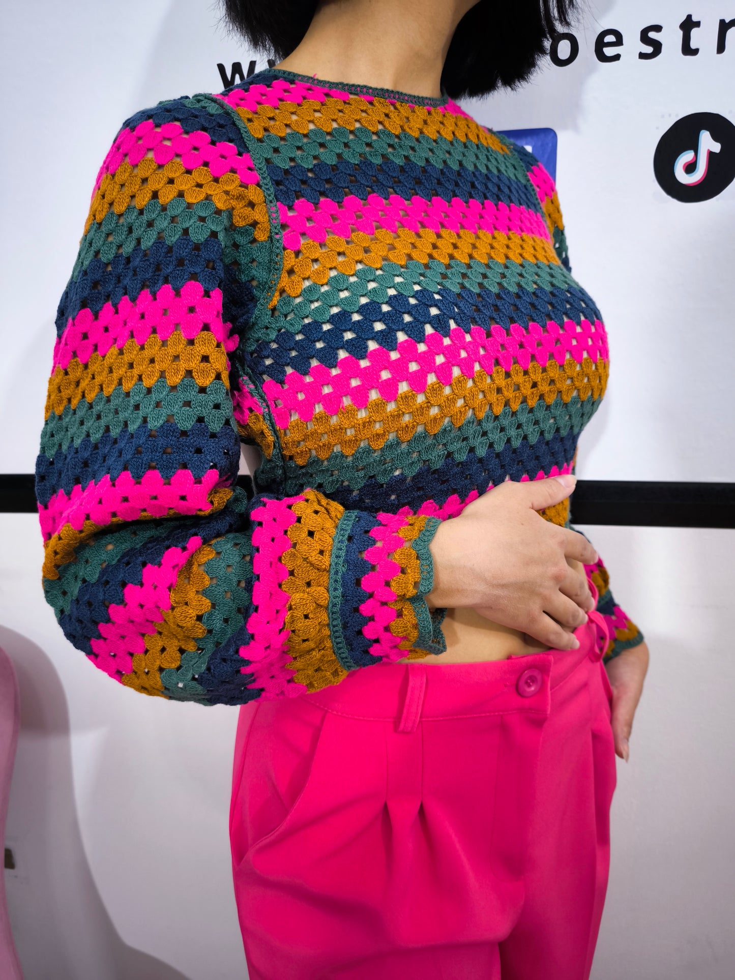 Maglia crochet multicolor a righe con schiena incrociata, abbinata a pantaloni fucsia e cintura dorata, indossata da una modella sorridente.