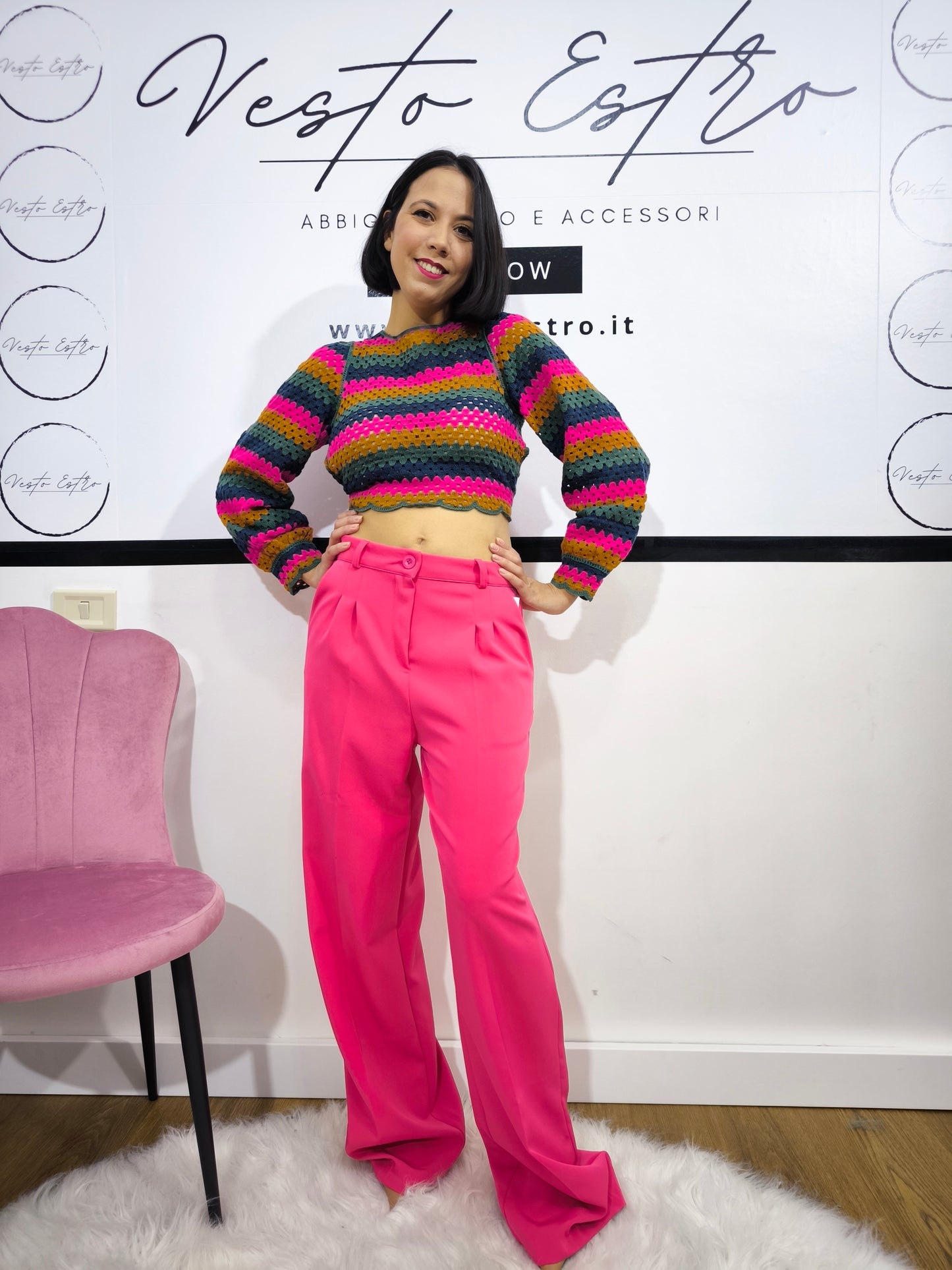 Maglia crochet multicolor a righe con schiena incrociata, abbinata a pantaloni fucsia e cintura dorata, indossata da una modella sorridente.