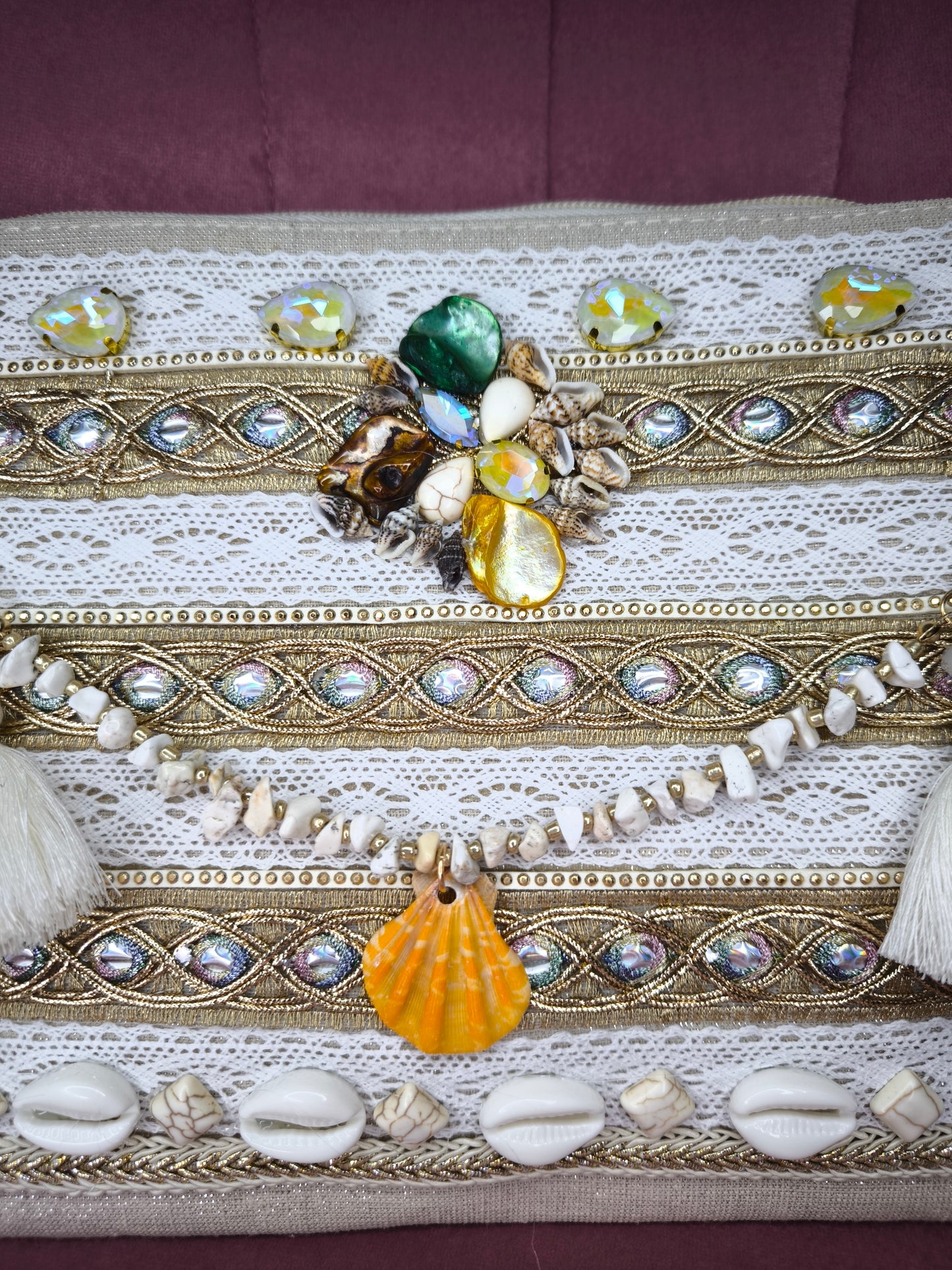 Pochette boho chic con tracolla e polsiera, decorata con pizzo, perline, conchiglie e pietre colorate, appoggiata su una sedia rosa.