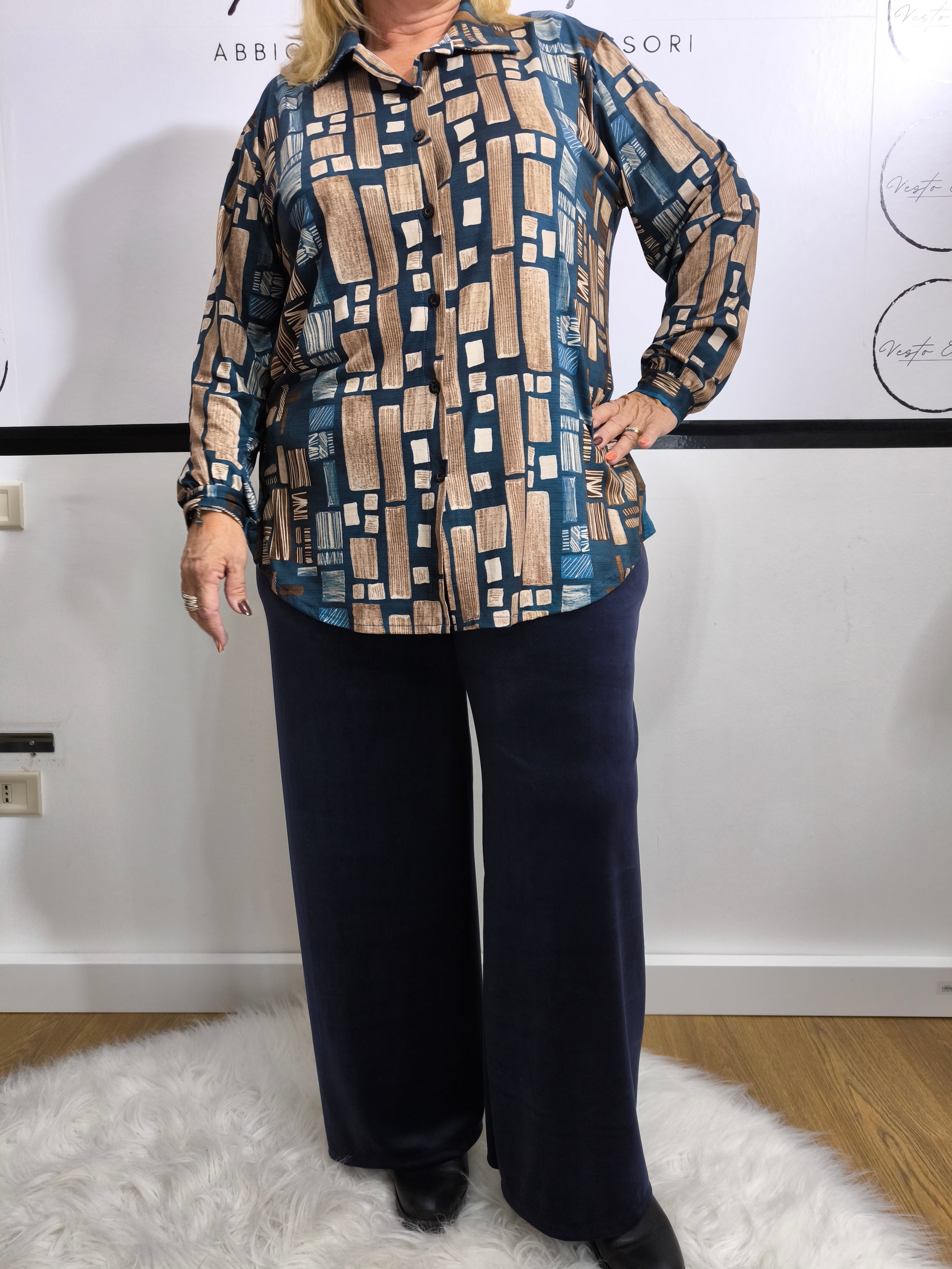 Donna che indossa una camicia in viscosa blu petrolio con stampa geometrica, affiancata dalla stessa camicia in tonalità nera e dorata su appendino.