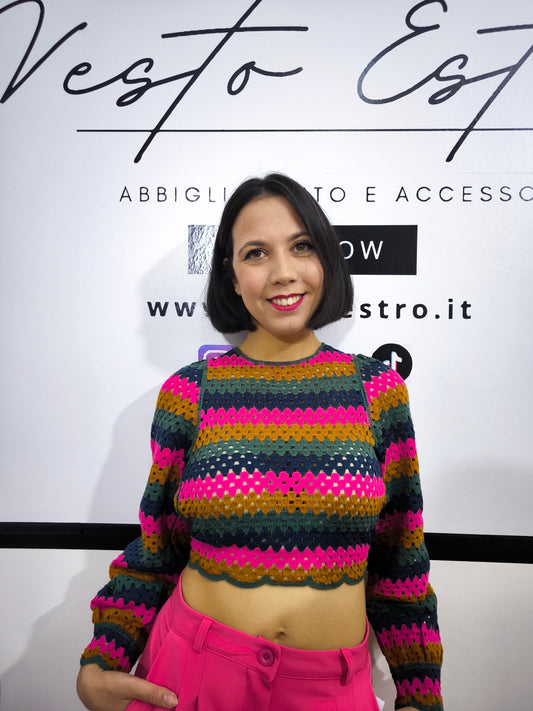 Maglia crochet multicolor a righe con schiena incrociata, abbinata a pantaloni fucsia e cintura dorata, indossata da una modella sorridente.