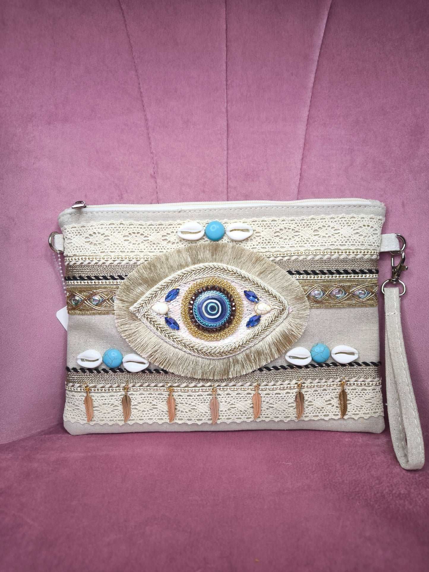 Pochette boho chic con occhio greco portafortuna, decorata con pizzo, conchiglie, piume dorate e dettagli turchesi, appoggiata su una sedia rosa.