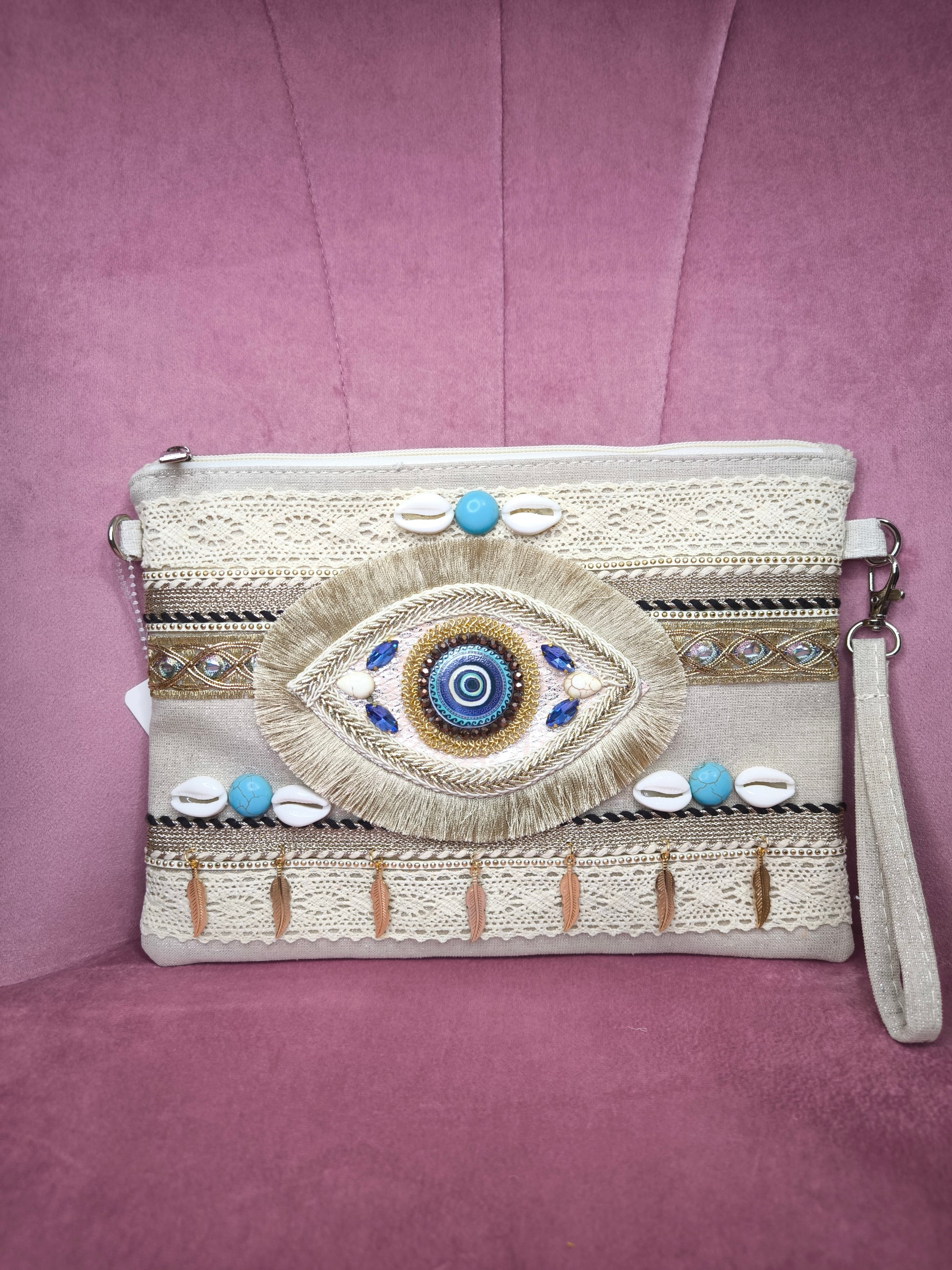 Pochette boho chic con occhio greco portafortuna, decorata con pizzo, conchiglie, piume dorate e dettagli turchesi, appoggiata su una sedia rosa.