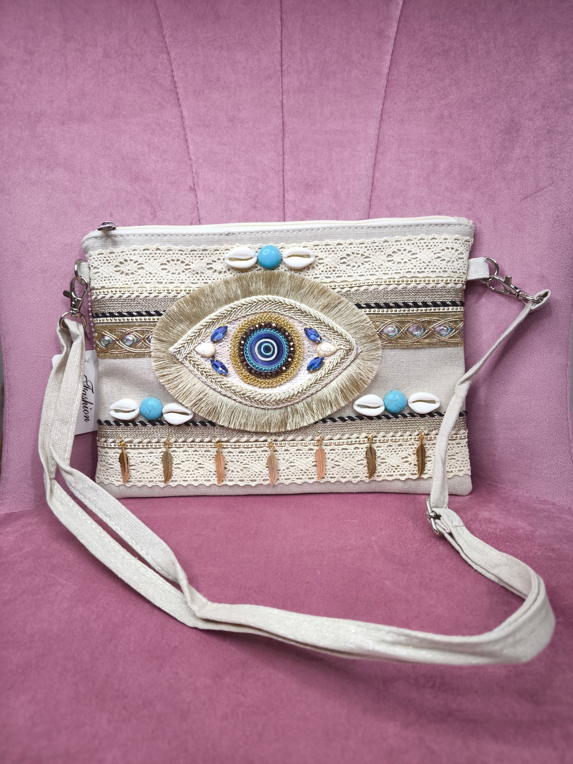 Pochette boho chic con occhio greco portafortuna, decorata con pizzo, conchiglie, piume dorate e dettagli turchesi, appoggiata su una sedia rosa.