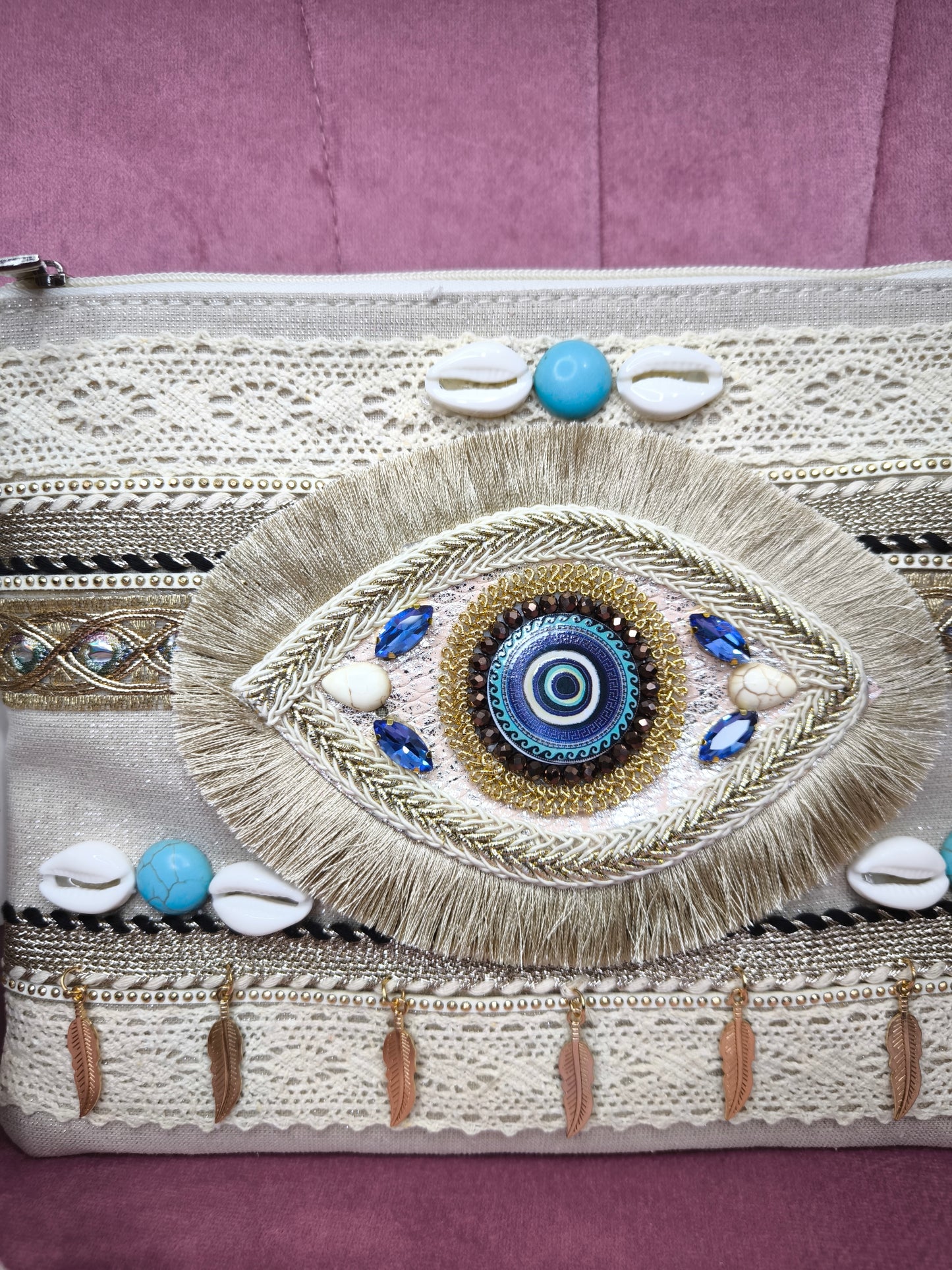 Pochette boho chic con occhio greco portafortuna, decorata con pizzo, conchiglie, piume dorate e dettagli turchesi, appoggiata su una sedia rosa.