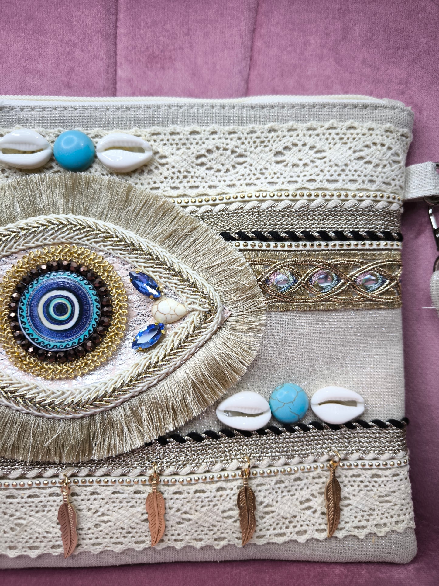 Pochette boho chic con occhio greco portafortuna, decorata con pizzo, conchiglie, piume dorate e dettagli turchesi, appoggiata su una sedia rosa.