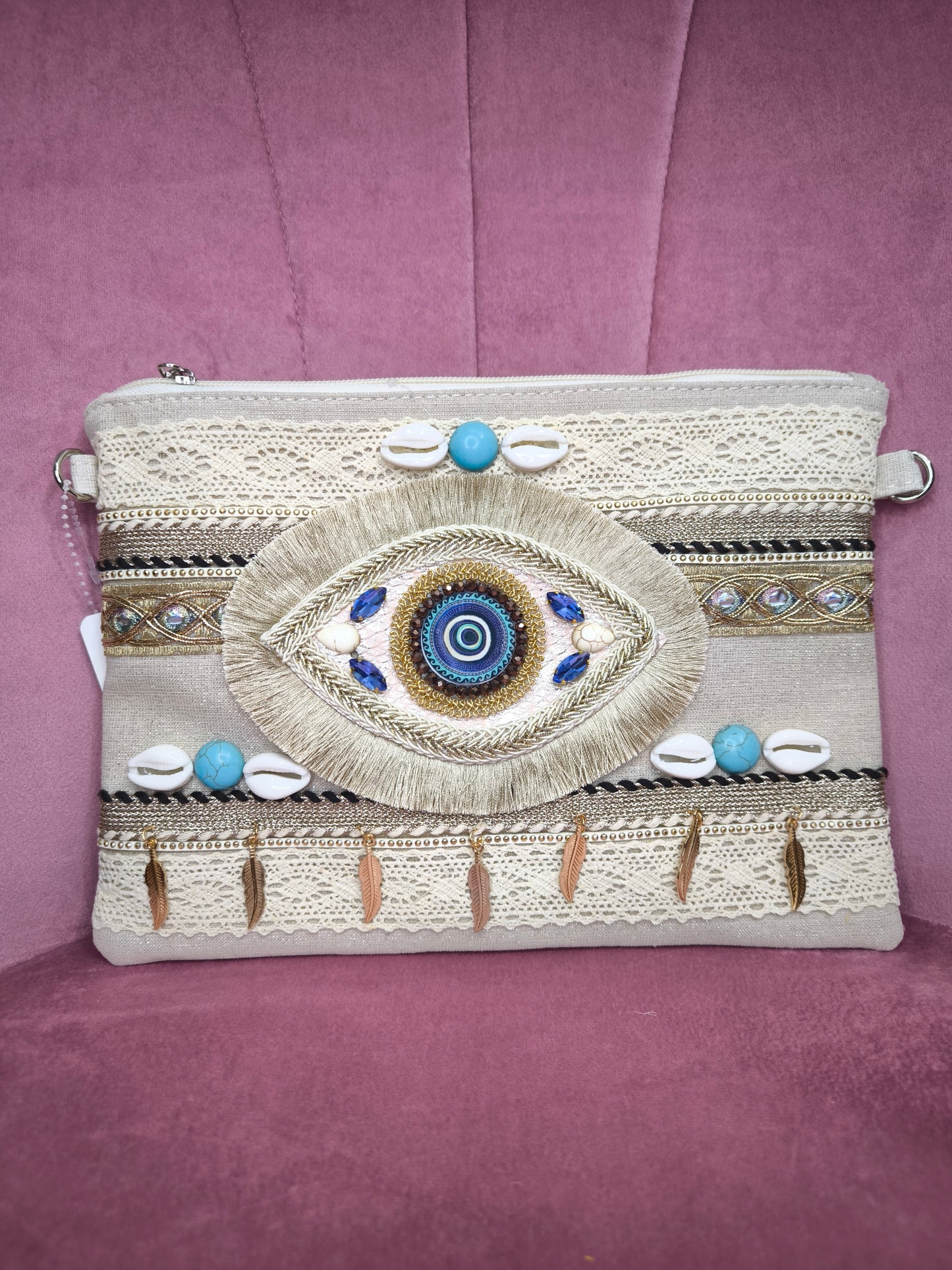 Pochette boho chic con occhio greco portafortuna, decorata con pizzo, conchiglie, piume dorate e dettagli turchesi, appoggiata su una sedia rosa.
