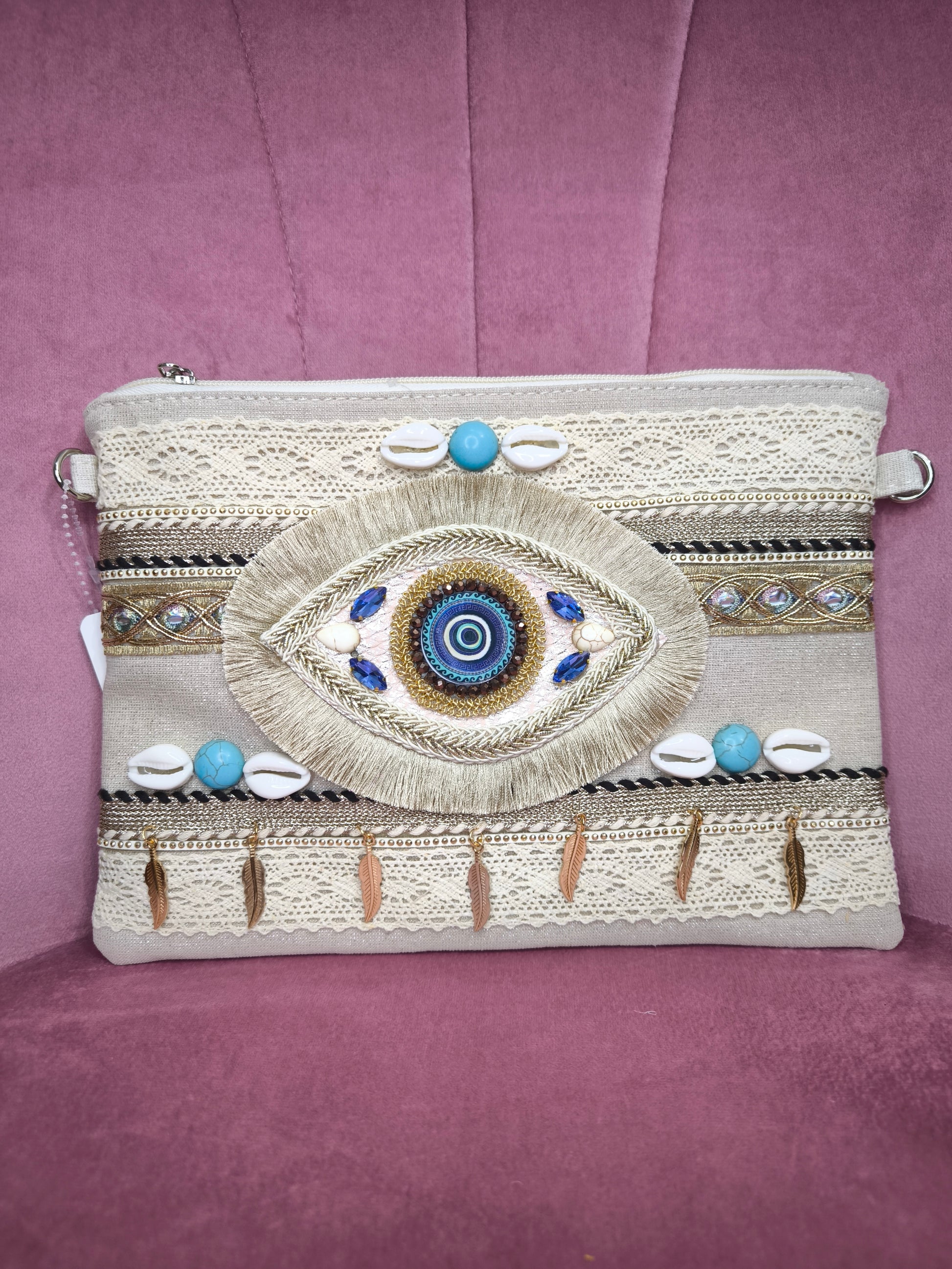 Pochette boho chic con occhio greco portafortuna, decorata con pizzo, conchiglie, piume dorate e dettagli turchesi, appoggiata su una sedia rosa.