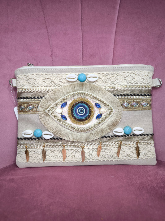 Pochette boho chic con occhio greco portafortuna, decorata con pizzo, conchiglie, piume dorate e dettagli turchesi, appoggiata su una sedia rosa.