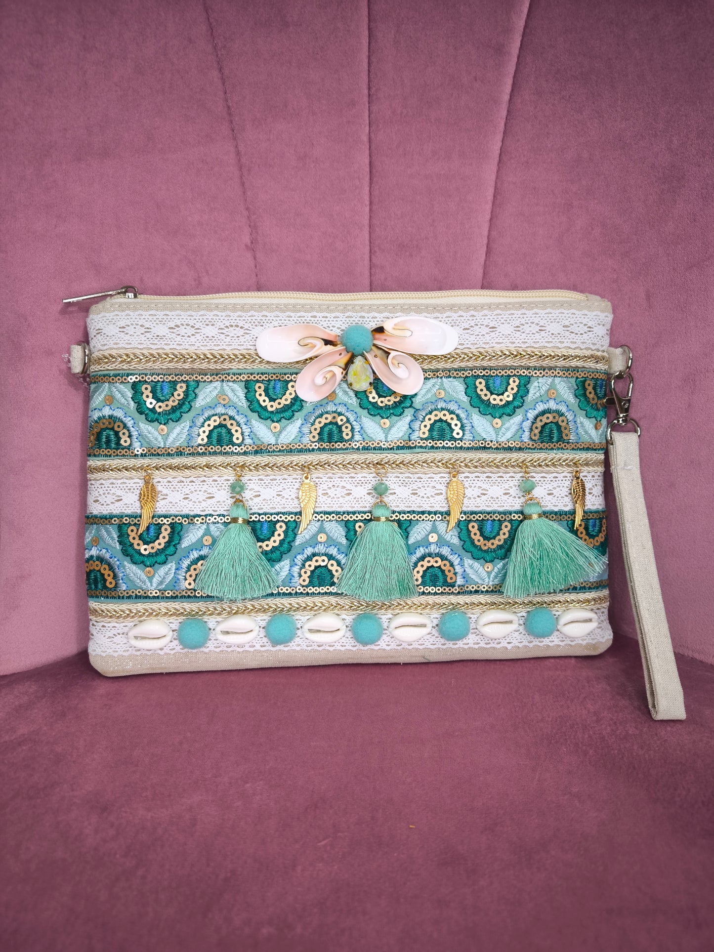 Pochette boho chic con tracolla e polsiera, decorata con pizzo, nappine verde acqua, conchiglie e dettagli dorati, appoggiata su una sedia rosa.