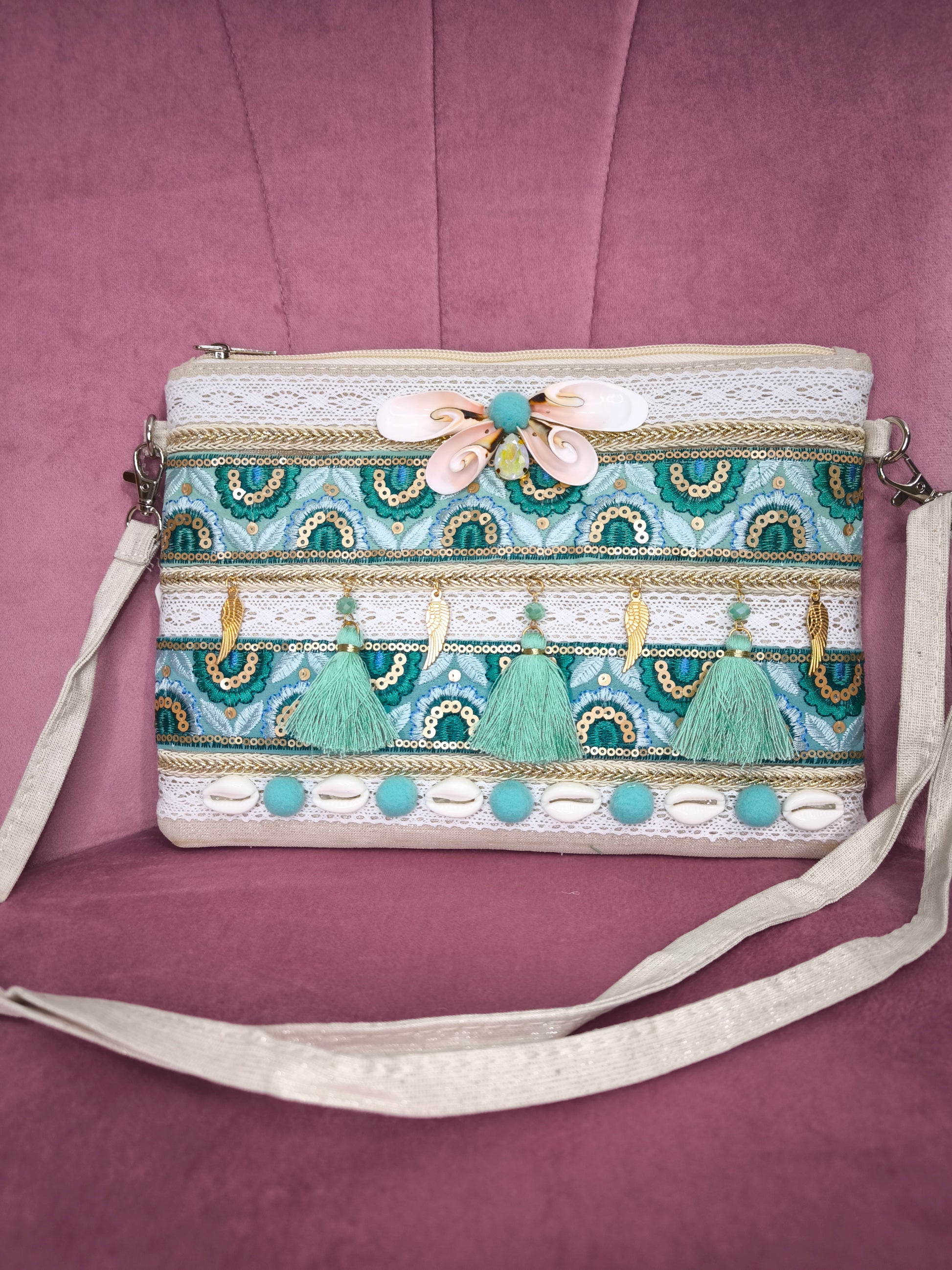 Pochette boho chic con tracolla e polsiera, decorata con pizzo, nappine verde acqua, conchiglie e dettagli dorati, appoggiata su una sedia rosa.