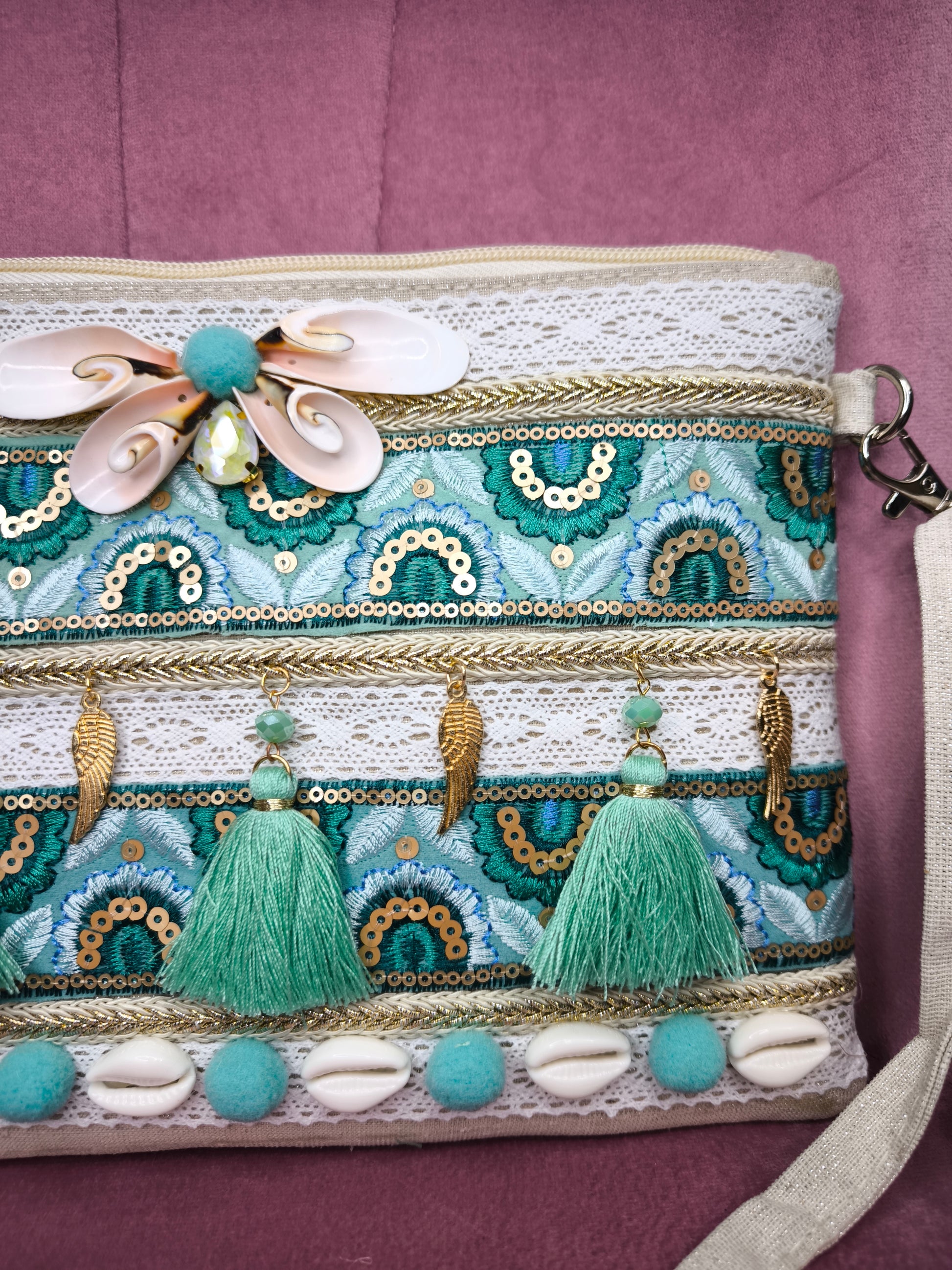 Pochette boho chic con tracolla e polsiera, decorata con pizzo, nappine verde acqua, conchiglie e dettagli dorati, appoggiata su una sedia rosa.