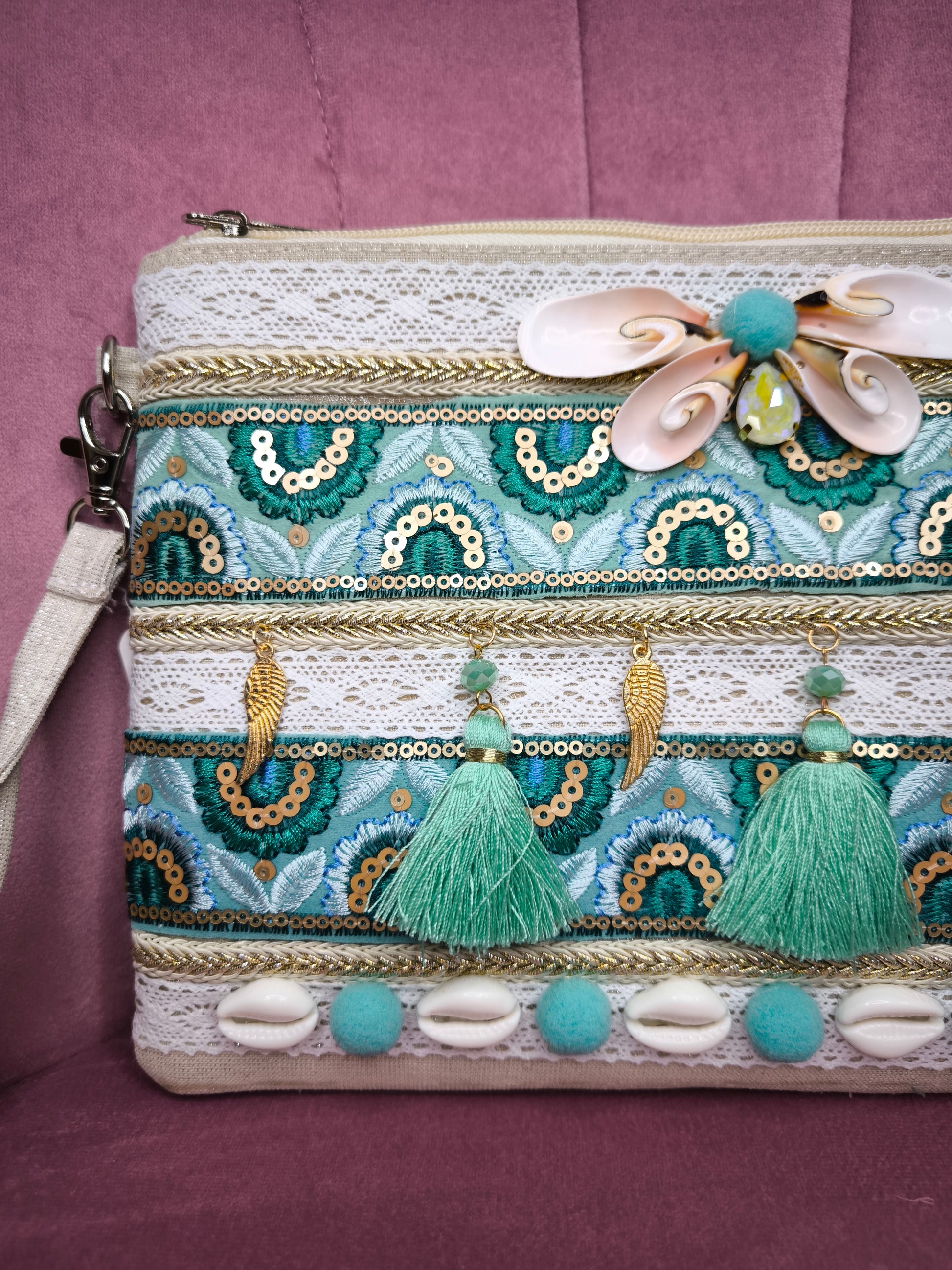 Pochette boho chic con tracolla e polsiera, decorata con pizzo, nappine verde acqua, conchiglie e dettagli dorati, appoggiata su una sedia rosa.