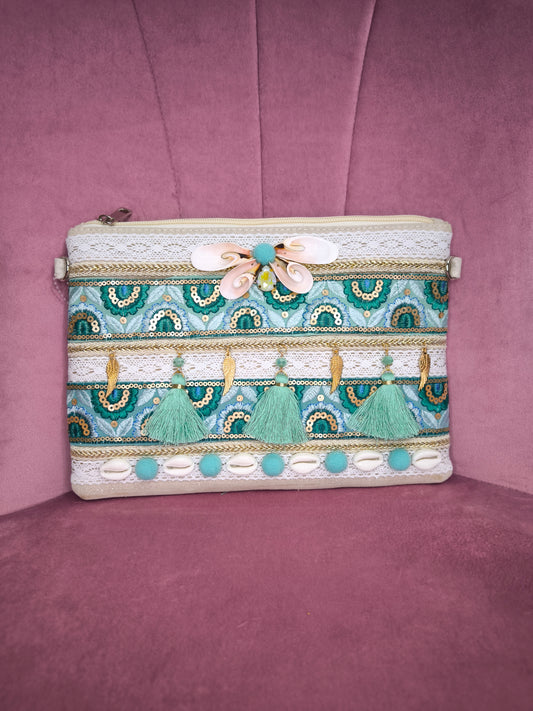 Pochette boho chic con tracolla e polsiera, decorata con pizzo, nappine verde acqua, conchiglie e dettagli dorati, appoggiata su una sedia rosa.
