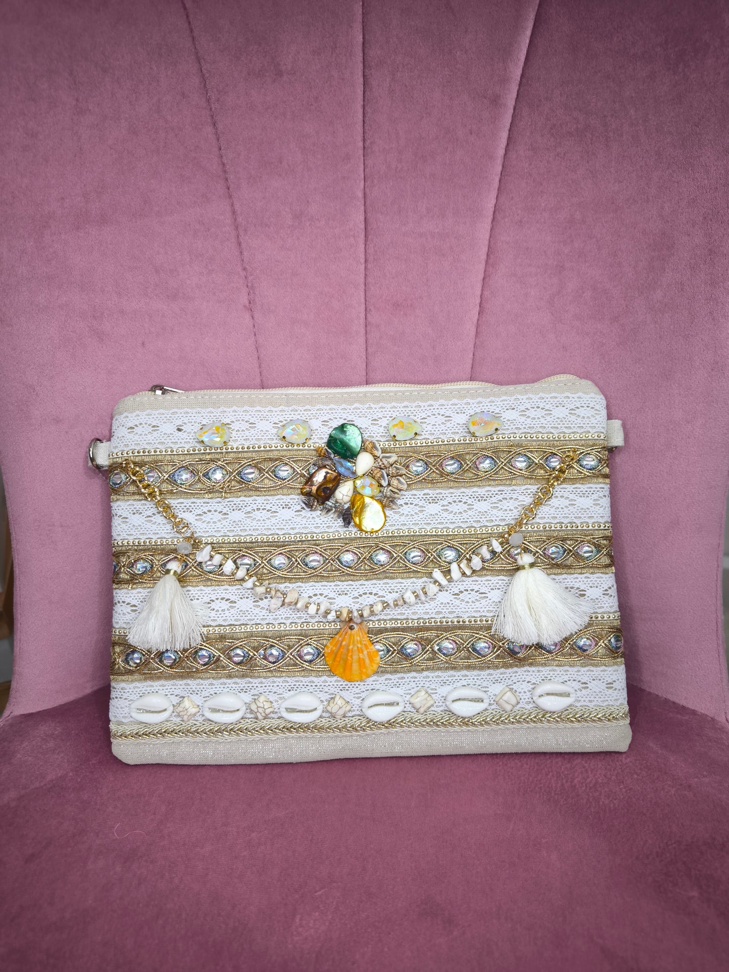 Pochette boho chic con tracolla e polsiera, decorata con pizzo, perline, conchiglie e pietre colorate, appoggiata su una sedia rosa.