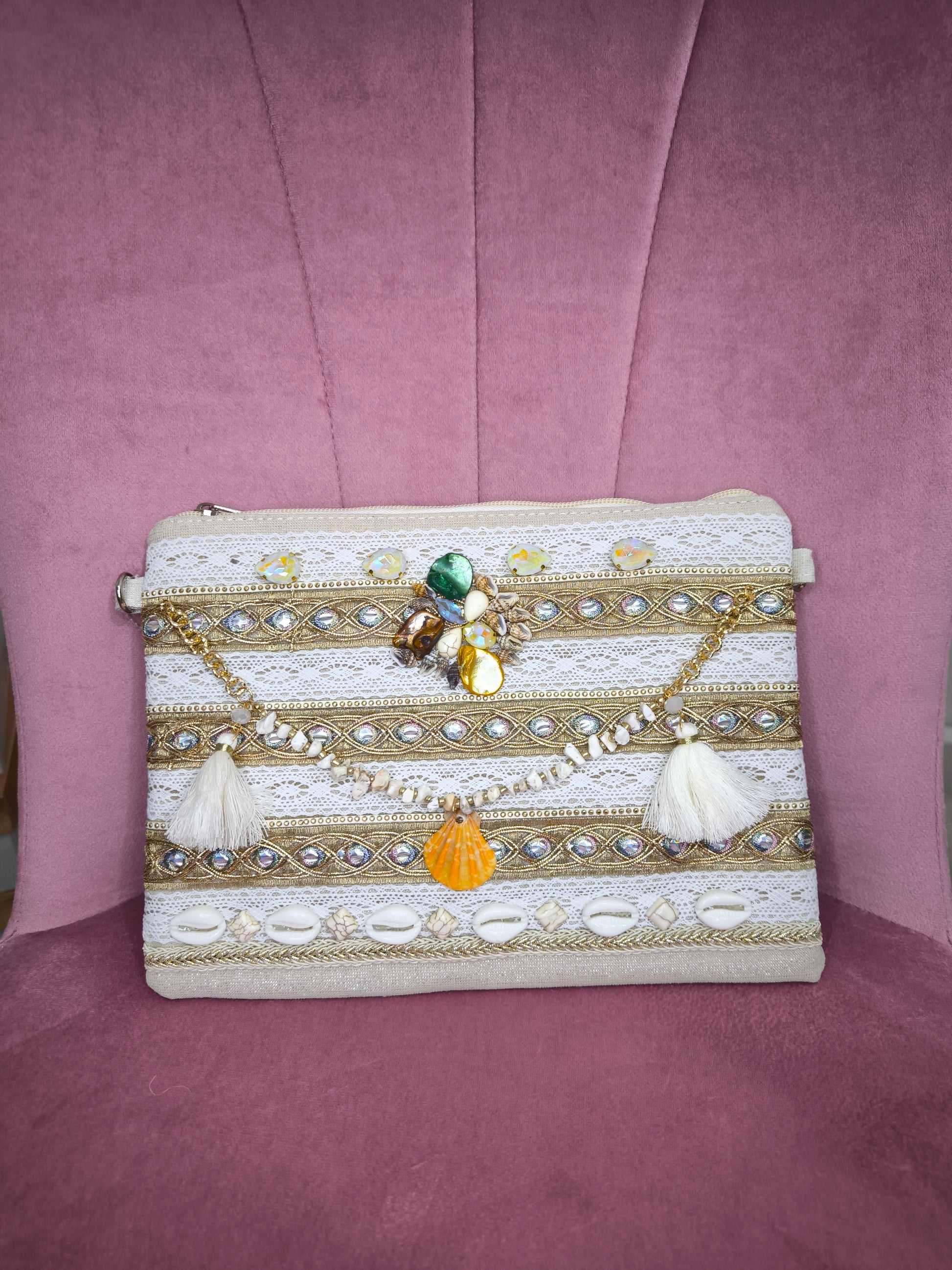 Pochette boho chic con tracolla e polsiera, decorata con pizzo, perline, conchiglie e pietre colorate, appoggiata su una sedia rosa.