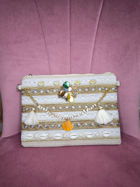 Pochette boho chic con tracolla e polsiera, decorata con pizzo, perline, conchiglie e pietre colorate, appoggiata su una sedia rosa.