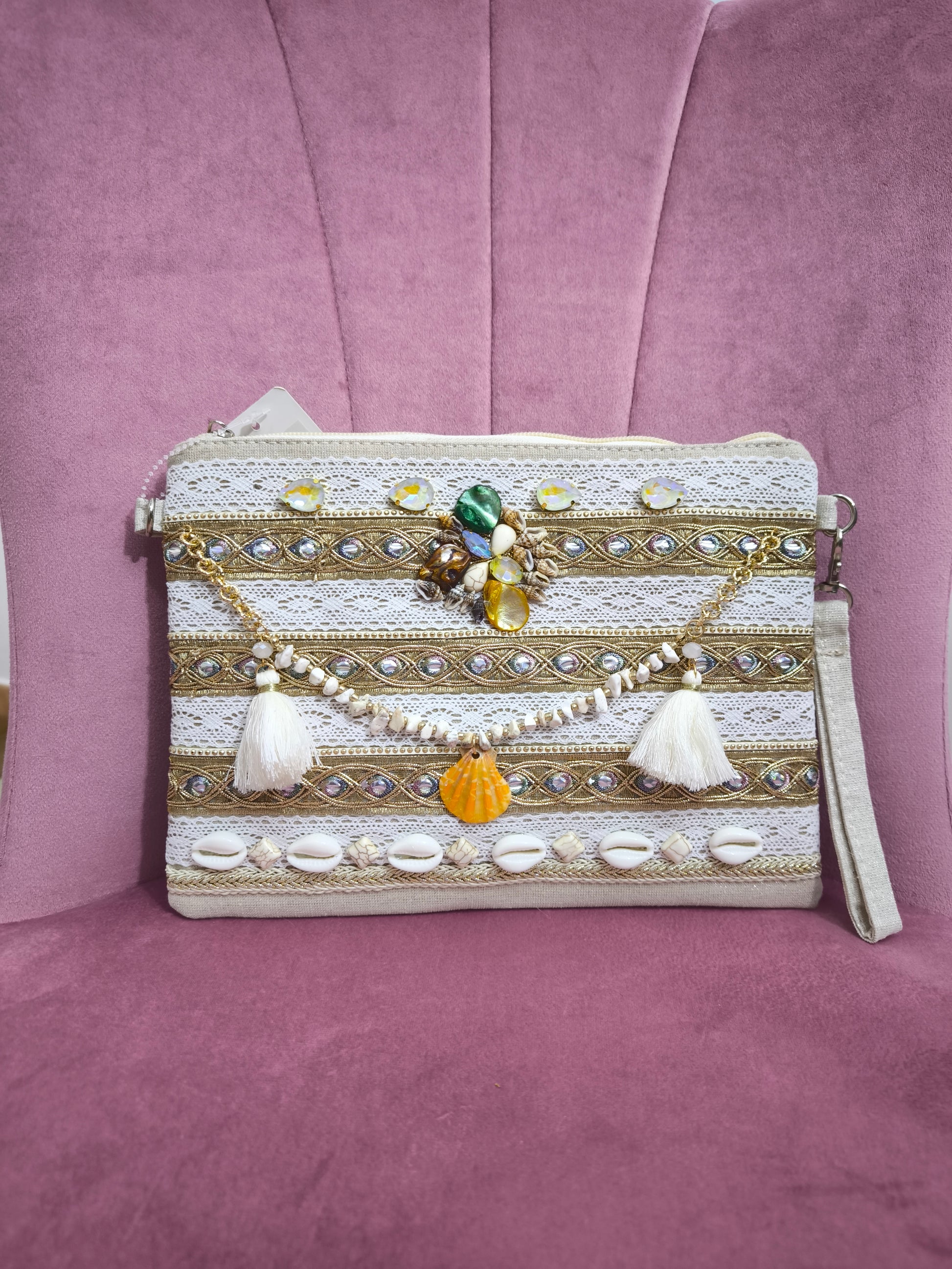 Pochette boho chic con tracolla e polsiera, decorata con pizzo, perline, conchiglie e pietre colorate, appoggiata su una sedia rosa.