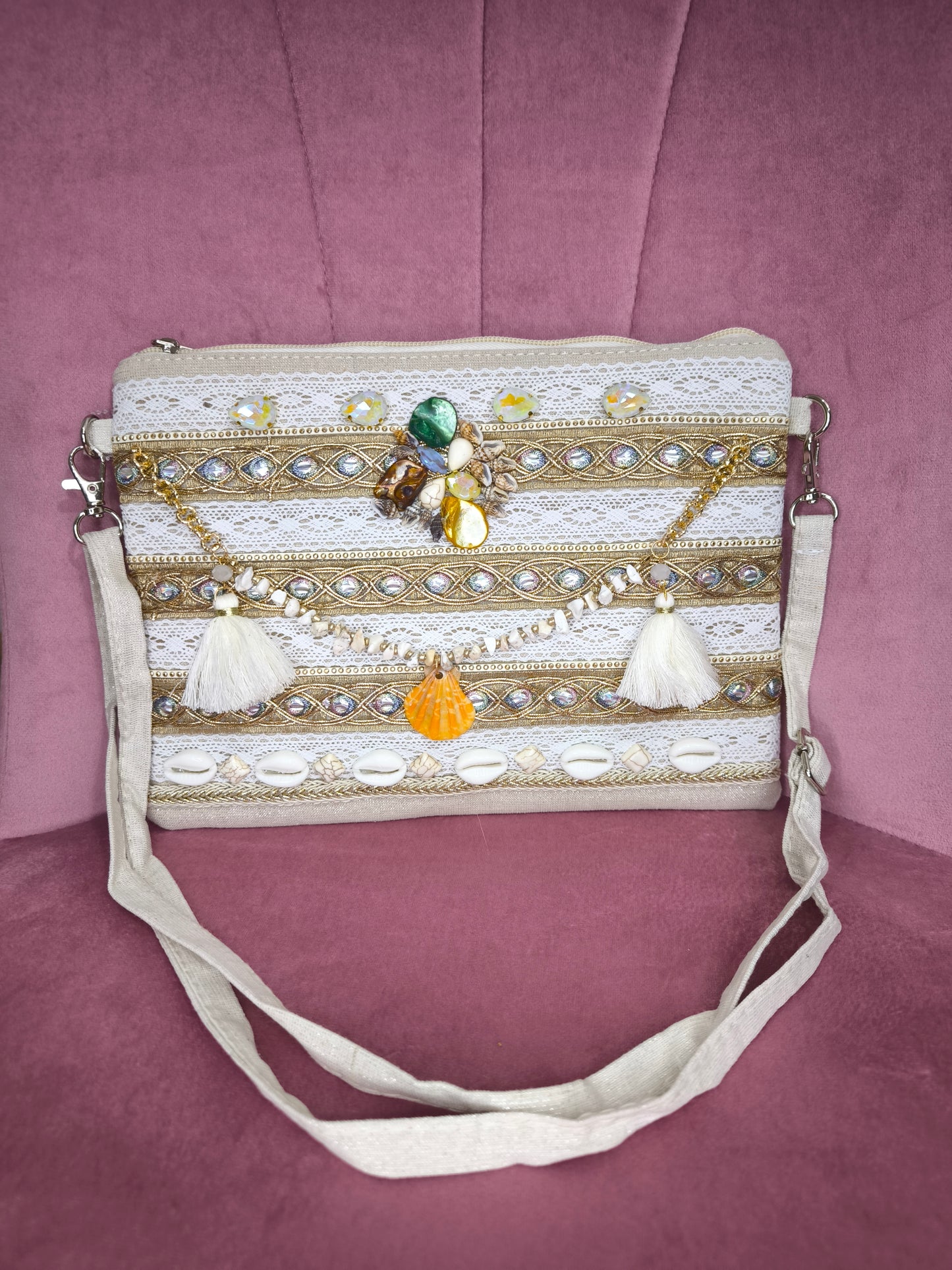 Pochette boho chic con tracolla e polsiera, decorata con pizzo, perline, conchiglie e pietre colorate, appoggiata su una sedia rosa.