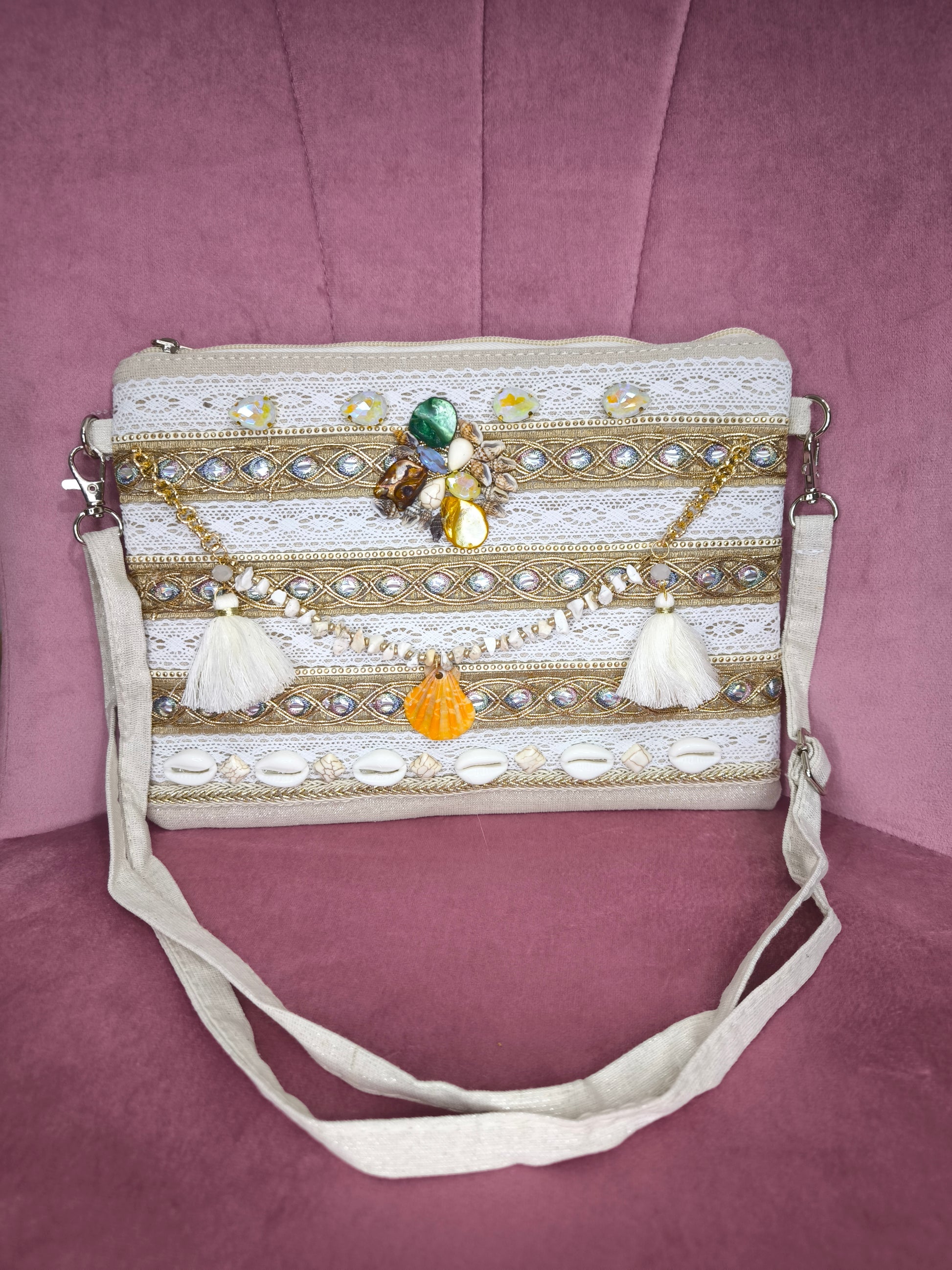 Pochette boho chic con tracolla e polsiera, decorata con pizzo, perline, conchiglie e pietre colorate, appoggiata su una sedia rosa.