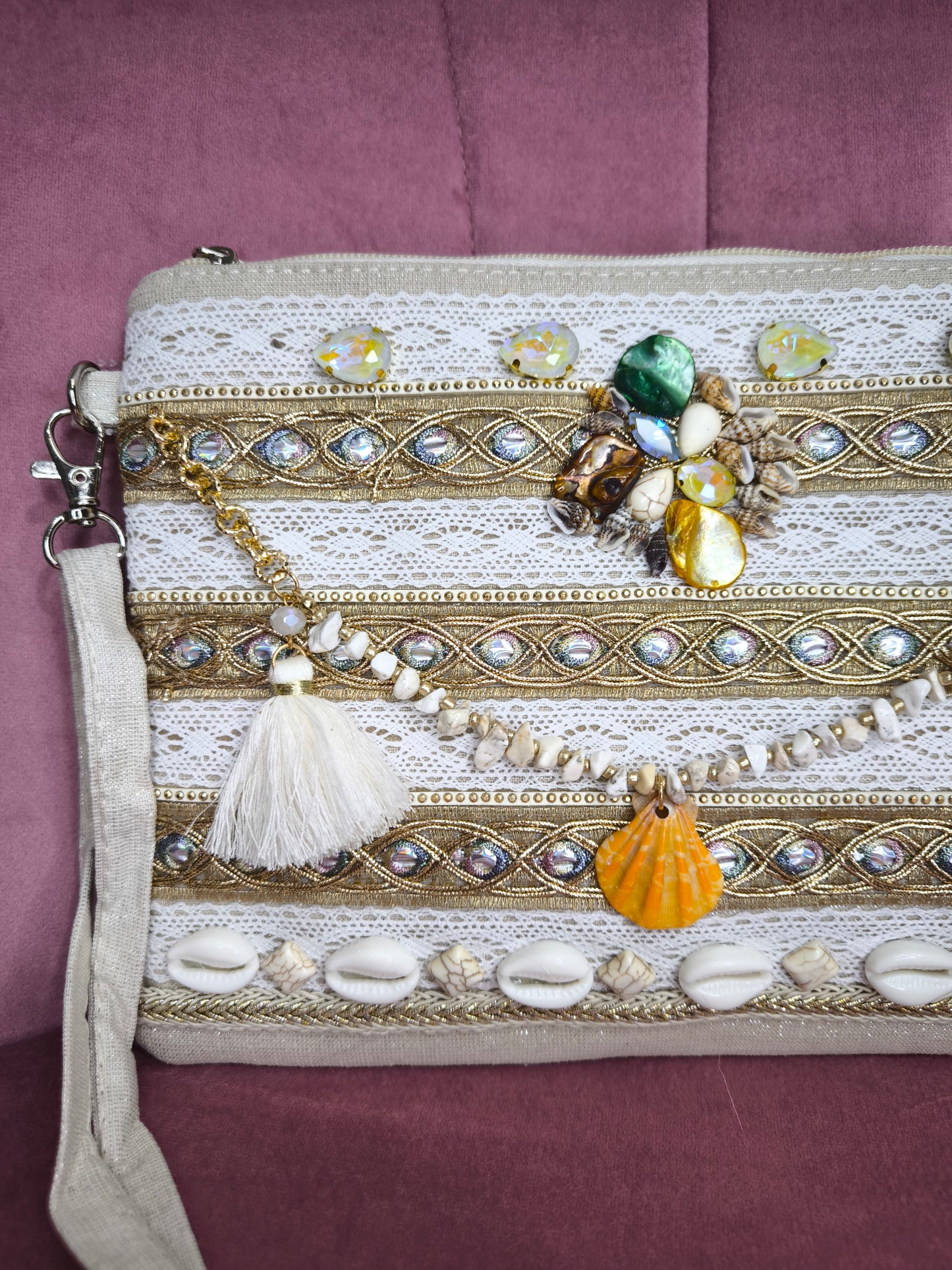 Pochette boho chic con tracolla e polsiera, decorata con pizzo, perline, conchiglie e pietre colorate, appoggiata su una sedia rosa.
