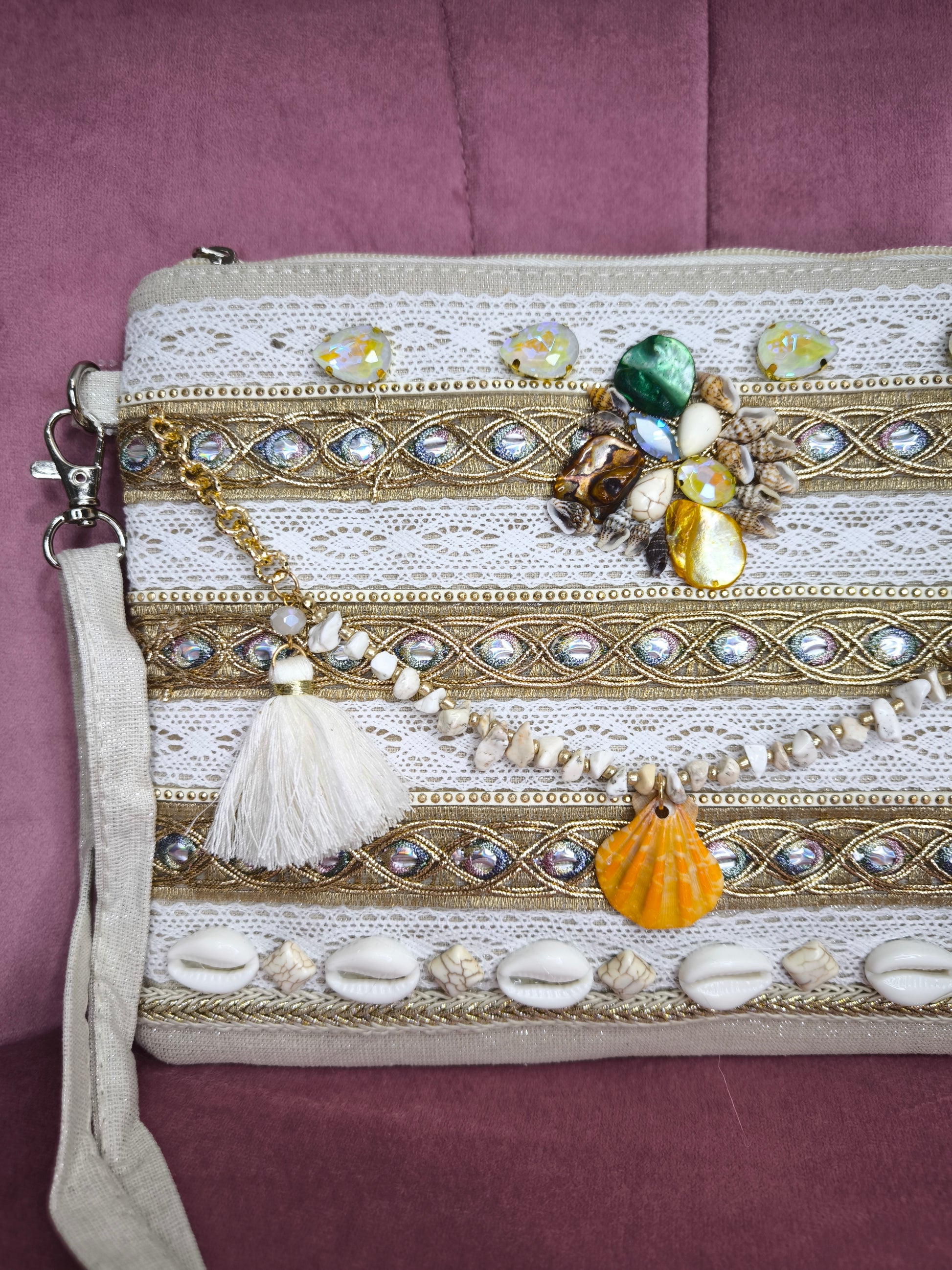 Pochette boho chic con tracolla e polsiera, decorata con pizzo, perline, conchiglie e pietre colorate, appoggiata su una sedia rosa.