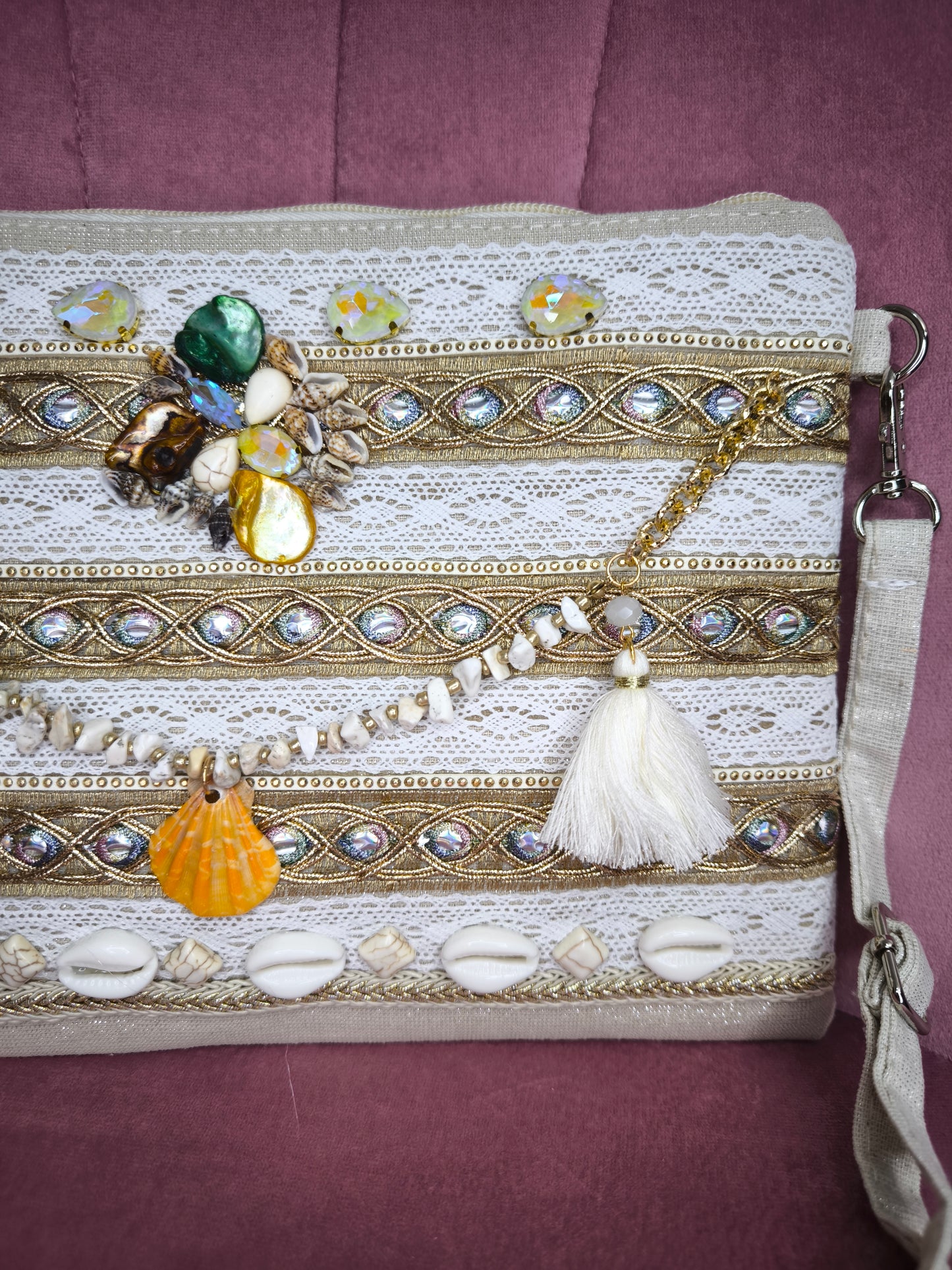 Pochette boho chic con tracolla e polsiera, decorata con pizzo, perline, conchiglie e pietre colorate, appoggiata su una sedia rosa.