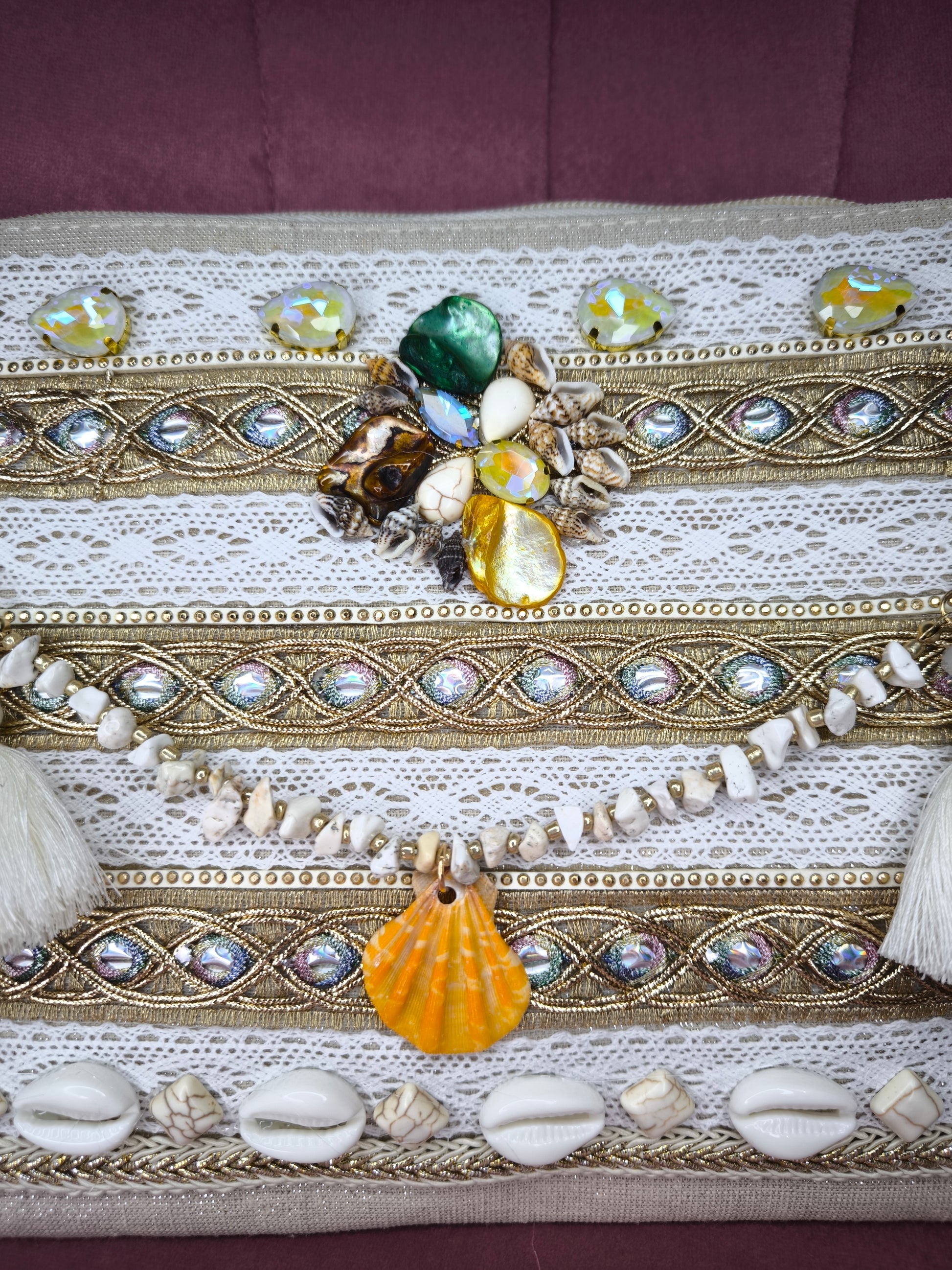 Pochette boho chic con tracolla e polsiera, decorata con pizzo, perline, conchiglie e pietre colorate, appoggiata su una sedia rosa.